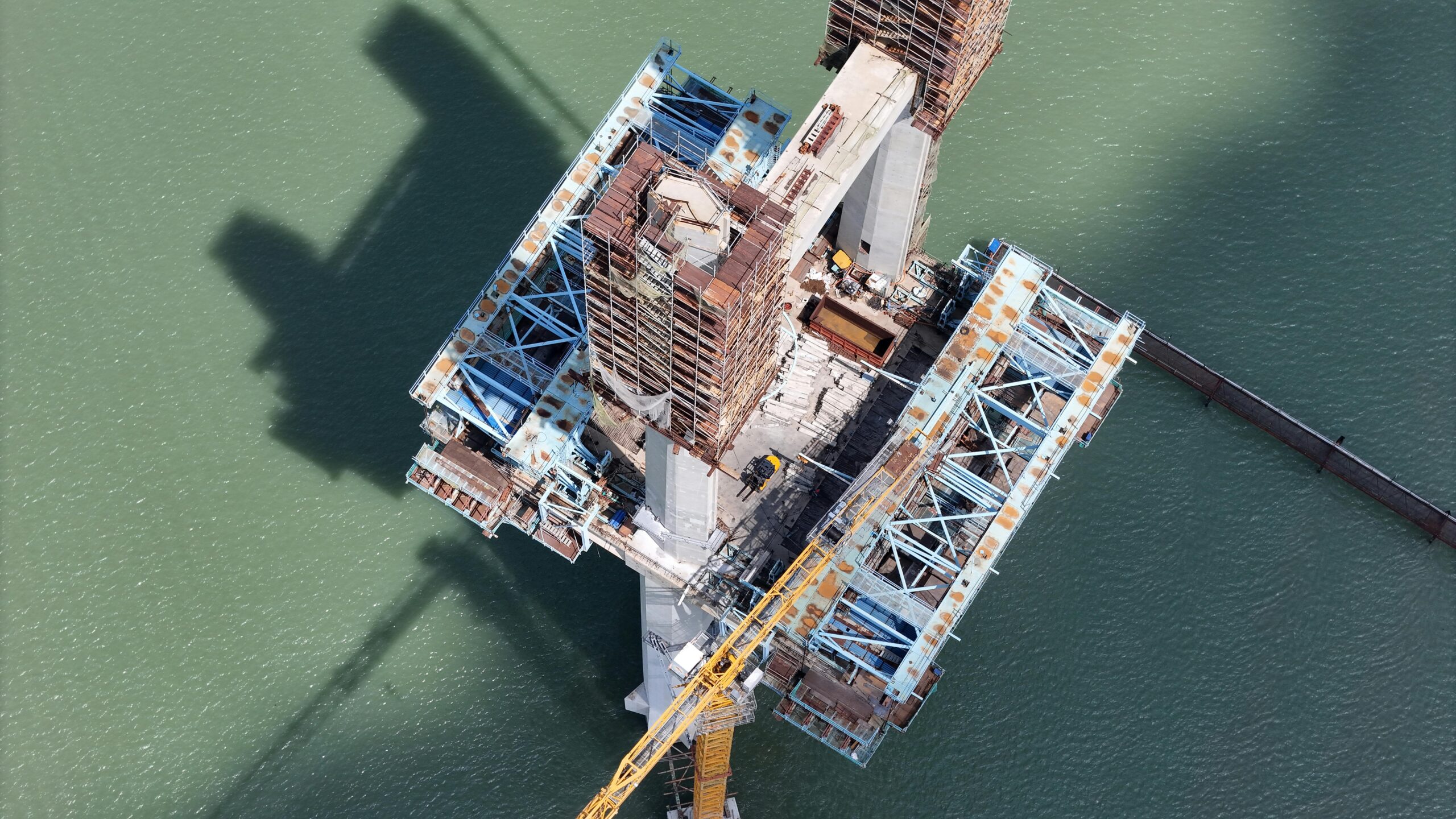 Aerial perspective of an unfinished bridge structure over a green river, showcasing industrial construction.